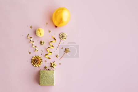 Photo for Birthday objects exploding from a gold sparkly gift box, overhead view, against a pink background - Royalty Free Image