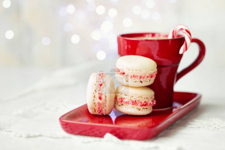 Foto de Macarons de Navidad decorados con bastones de caramelo triturados junto a la taza roja de chocolate caliente - Imagen libre de derechos