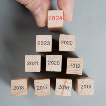 Photo for Photo of wooden blocks in a triangle shape with gray numbers and one of them standing out hold by a human hand red number 2024 imprinted on wooden surface. New Year concept. - Royalty Free Image