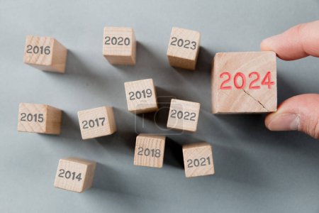 Photo for Photo of wooden block group with gray numbers of passed years and one of them hold by a human hand with red number 2024 imprinted on wooden surface. New Year, Strategy, Future and Standing Out concept. - Royalty Free Image