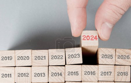 Photo for Photo of wooden block rows with gray numbers and one of them hold by a human hand with red number 2024 imprinted on wooden surface. Gray backgound. New Year, Strategy, Vision and Standing Out concept. - Royalty Free Image