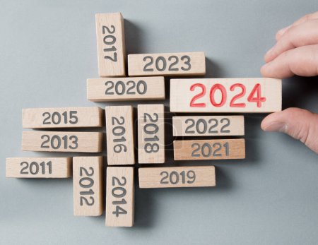 Photo for Photo of wooden blocks with gray numbers and one of them standing out and hold by a human hand with red number 2024 imprinted on wooden surface. Gray Background. New Year, Future, Vision and Standing Out concept. - Royalty Free Image