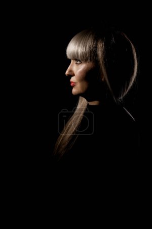 Beautiful girl with blond bangs fringe against black background. Abstract sidelit portrait.
