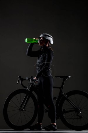 Photo for Girl posing on roadbike. Drinking from green cycling water bottle. White protective helmet. Side lit cyclist against dark background. - Royalty Free Image