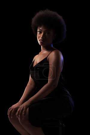 Téléchargez les photos : Studio portrait of elegant african american lady with curly hair afro hairstyle against black background. Girl in black dress. - en image libre de droit