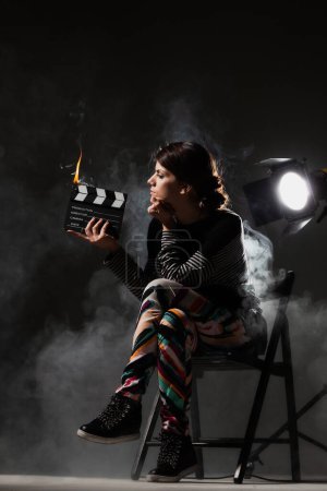 Photo for Female movie director on set with smoke background. Girl holding clapperboard on fire. - Royalty Free Image