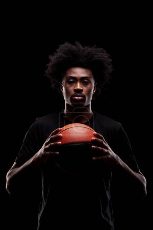 Foto de Jugador de baloncesto sosteniendo una pelota contra fondo negro. Hombre afroamericano concentrado serio. - Imagen libre de derechos
