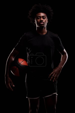 Foto de Basketball player holding a ball. Young african american sports man against black background. - Imagen libre de derechos