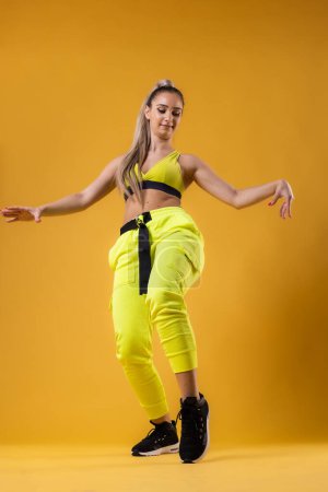Foto de Hermosa chica en traje amarillo bailando zumba. Instructor de baile feliz sobre fondo amarillo oscuro o naranja. - Imagen libre de derechos