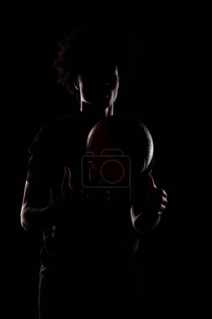 Foto de Side lit silhouette of a basketball player. African American man holding basket ball posing against black background. - Imagen libre de derechos