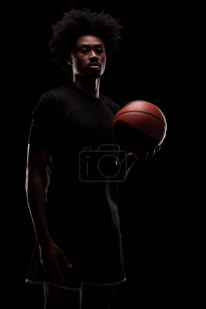 Foto de Jugador de baloncesto sosteniendo una pelota contra fondo negro. Hombre afroamericano concentrado serio. - Imagen libre de derechos