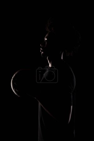 Foto de Side lit silhouette of a basketball player. African American man holding basket ball posing against black background. - Imagen libre de derechos