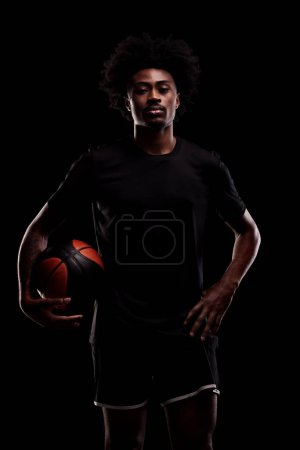 Foto de Basketball player holding a ball. Young african american sports man against black background. - Imagen libre de derechos