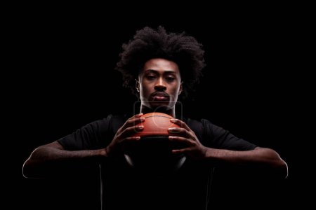 Basketball player holding a ball against black background. Serious concentrated african american man.
