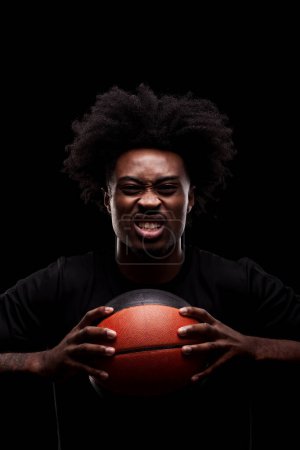 Basketball player holding a ball against black background and screaming. African american man with serious angry powerful face expression.