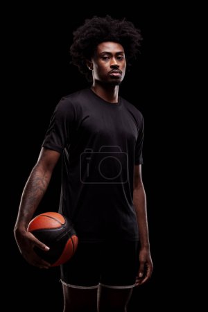 Basketball player holding a ball. Young african american sports man against black background.