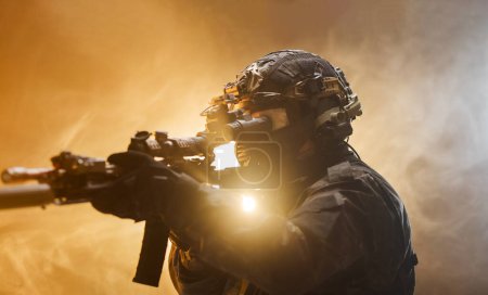 Focused man holding a gun in his hand in a serious manner. Soldier in full professional army outfit against fog background with flashing backlight.