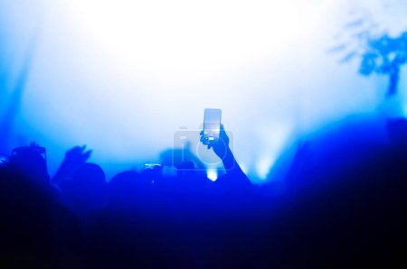 Photo for Silhouette of a hand holding a smartphone capturing a moment during a night concert lit by stage lights - Royalty Free Image