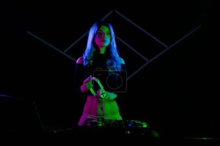 A woman with vibrant blue hair is DJing on a turntable. Holding headphones and looking at camera. Sidelit with colorful neon lights