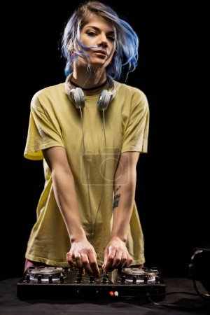 Chica con auriculares se centra en la mezcla de música en una caja de resonancia. DJ femenino con pelo azul y camiseta amarilla.