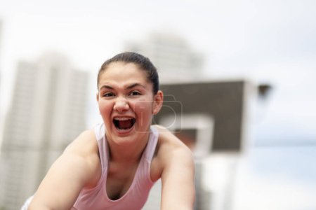Basketball player with angry screaming face expression. Girl posing with ball in hands. Outdoor sports field with urban background.