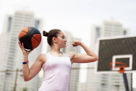 Basketball player portrait. Girl posing and showing muscles. Outdoor sports field with urban background.