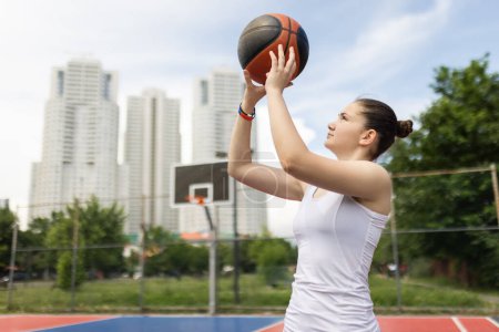 Young girl playing basketball. Holding ball in hands. Outdoor sports field with urban background.
