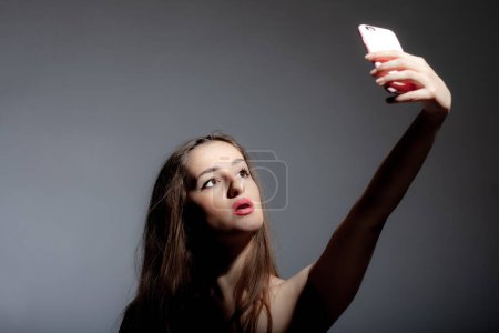 Beautiful girl looking at phone taking seplfy photo studio portrait against gray background.