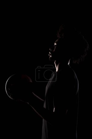 Side lit silhouette of a basketball player. African American man holding basket ball posing against black background.