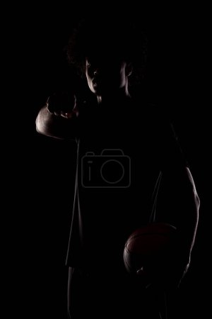 Basketball player holding a ball and making a fist. Young african american sports man against black background.