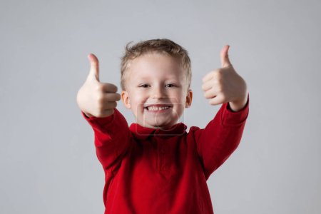 A young boy wearing a red shirt is smiling and giving a thumbs up gesture