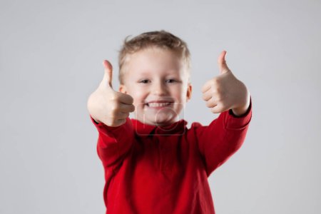 Un jeune garçon portant une chemise rouge sourit et fait un geste de pouce