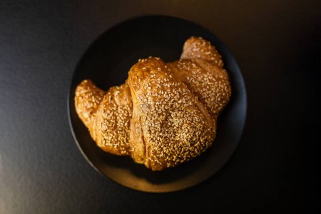 Dieses Bild fängt ein frisch gebackenes Croissant ein, großzügig mit Sesam überzogen, präsentiert auf einem dunklen, eleganten Teller, der an ein luxuriöses Frühstück oder einen Snack erinnert..
