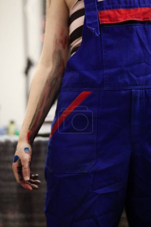 Close-up view of a female artist in a blue overalls, her hands smeared with vibrant paint colors, highlighting the messy and creative process of painting.