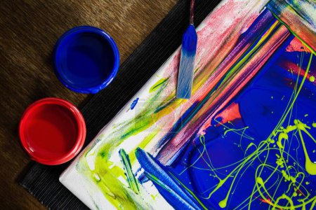 A close-up view of an abstract painting in progress featuring bright red and blue paints on canvas, highlighted by artist's brushes and containers.