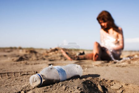 A thought-provoking scene showing a woman lamenting over pollution as she sits near a discarded plastic bottle on a sandy beach, symbolizing the devastating impact of human waste on natural environments.
