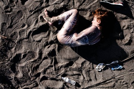 Vue aérienne d'une femme enveloppée de plastique transparent, allongée sur une plage couverte de boue entourée de déchets éparpillés, représentant la pollution et les problèmes environnementaux.
