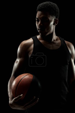 Basketball player holding a ball against black background. Serious concentrated african american man. Muscular person sidelit silhouette.