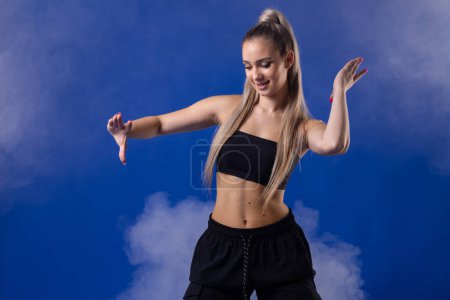 Foto de Hermosa chica rubia en traje negro bailando zumba contra niebla de humo y fondo azul. - Imagen libre de derechos
