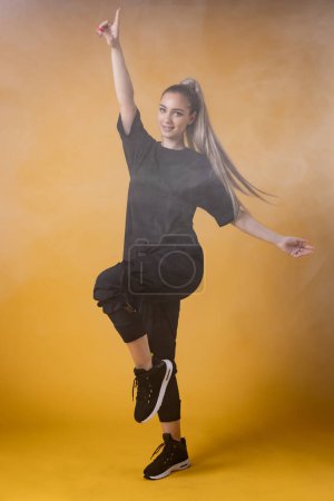 Foto de Hermosa chica rubia en camisa negra posando poses de baile contra el humo y la niebla de fondo amarillo. - Imagen libre de derechos