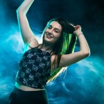 Happy young woman enjoying the moment while dancing energetically in smoky blue and green lights during a lively party.