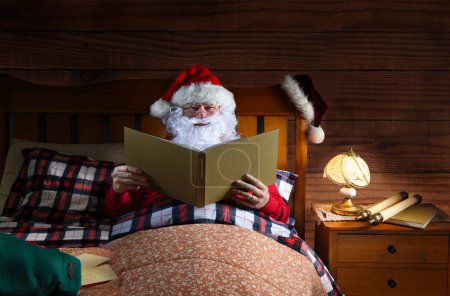 Photo for Santa Claus lying in his bed reading a large book with a sack of letters beside him. - Royalty Free Image