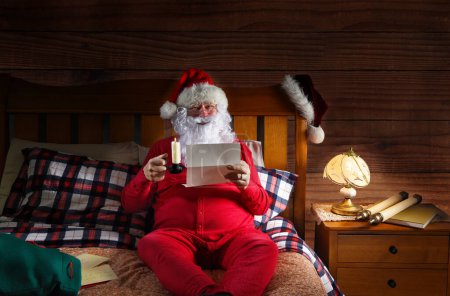 Photo for Santa Claus sitting on his bed wearing red long johns holding a candle and reading letters. - Royalty Free Image