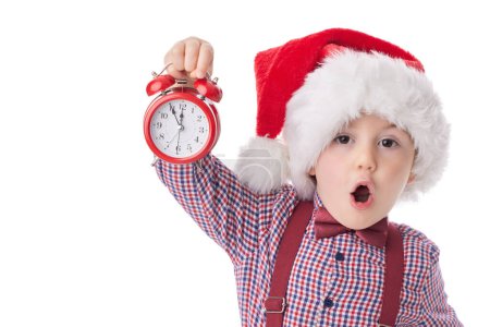 Photo for Christmas boy with red alarm clock, smiling little man in red Santa claus hat, tie and suspender posing on white background - Royalty Free Image