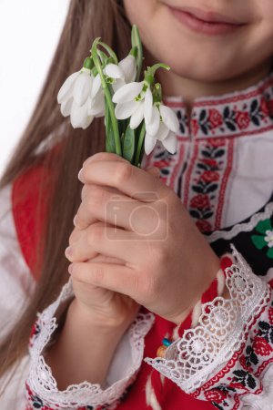 Photo for Bulgarian girl in traditional folklore costumes with snowdrop flowers and handcraft wool bracelet martenitsa symbol of Baba Marta, spring and Easter holiday,martisor - Royalty Free Image