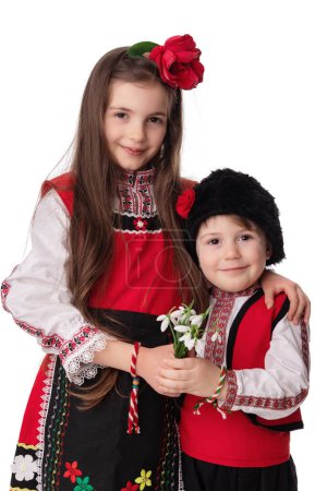 Photo for Bulgarian kids boy and girl in traditional folklore costumes with snowdrop flowers and handcraft wool bracelet martenitsa symbol of Baba Marta, spring and Easter, studio portrait.Martisor - Royalty Free Image