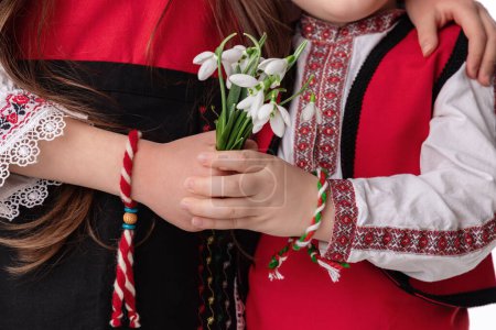 Photo for Bulgarian kids boy and girl in traditional folklore costumes with snowdrop flowers and handcraft wool bracelet martenitsa symbol of Baba Marta, spring and Easter, studio portrait,martisor - Royalty Free Image