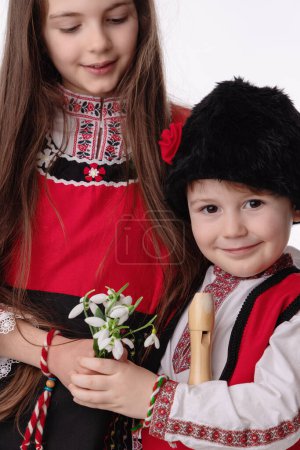 Bulgarian kids boy and girl in traditional folklore costumes with spring flowers snowdrop and handcraft wool bracelet martenitsa symbol of Baba Marta