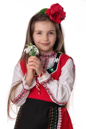 Chica búlgara en trajes folclóricos tradicionales con flores de primavera nevada y artesanía pulsera de lana martenitsa símbolo de Baba Marta