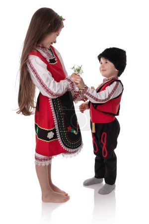 Photo for Bulgarian kids boy and girl in traditional folklore costumes with spring flowers snowdrop and handcraft wool bracelet martenitsa symbol of Baba Marta - Royalty Free Image